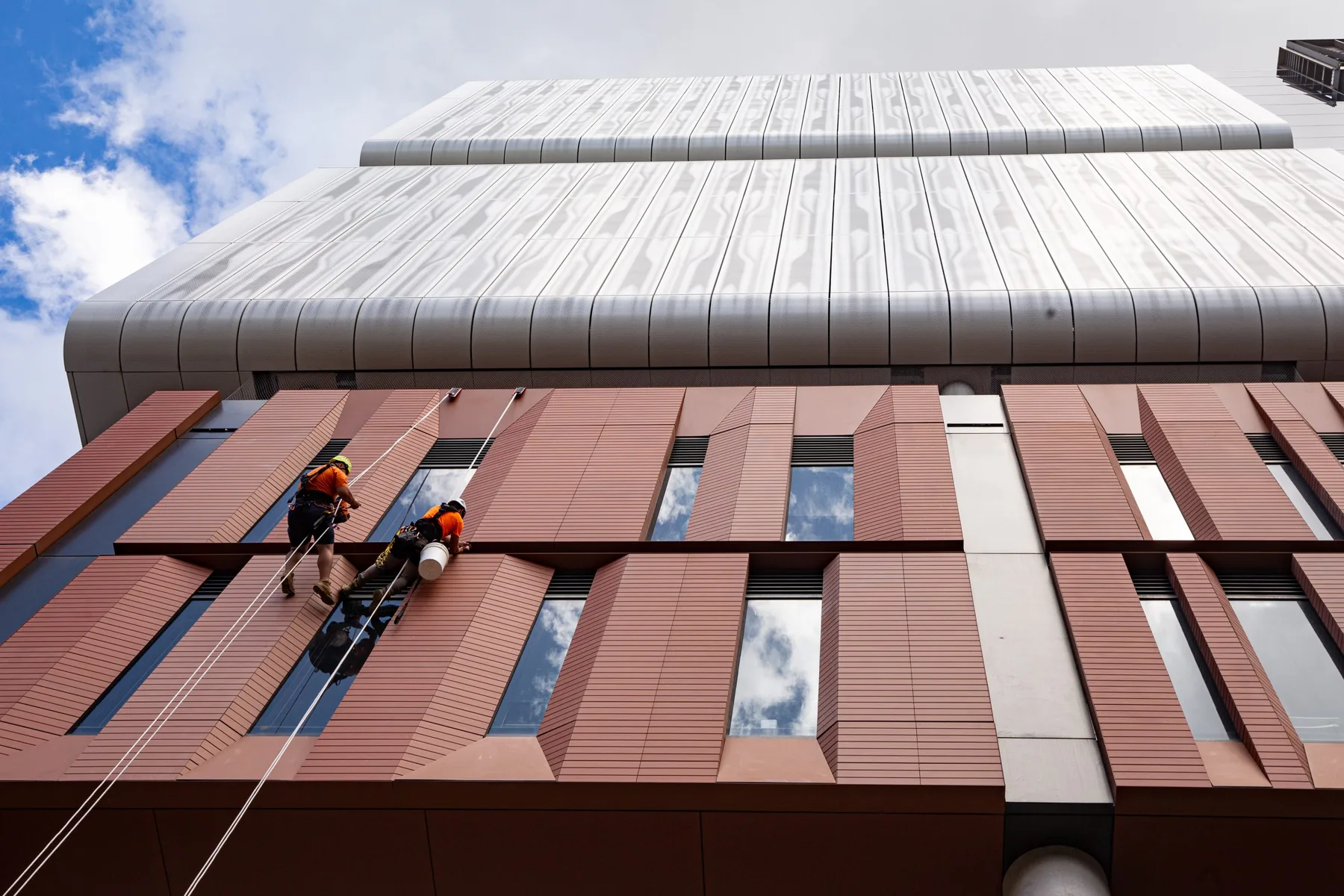 Two rope access technicians abseiling down a red-clad building facade