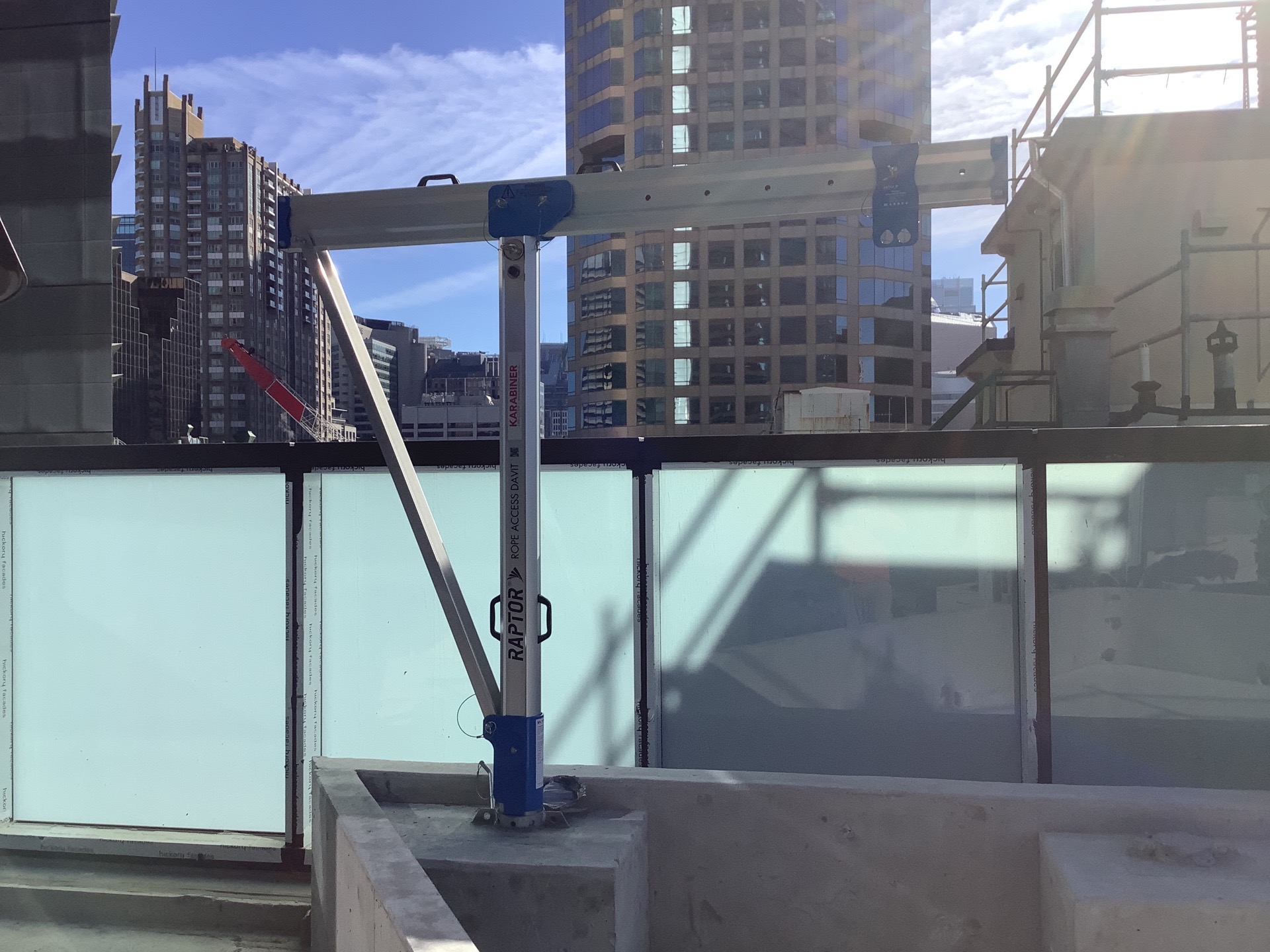 Davit arm set in a fixed base on a concrete rooftop upstand behind a glass balustrade, with Sydney CBD towers behind