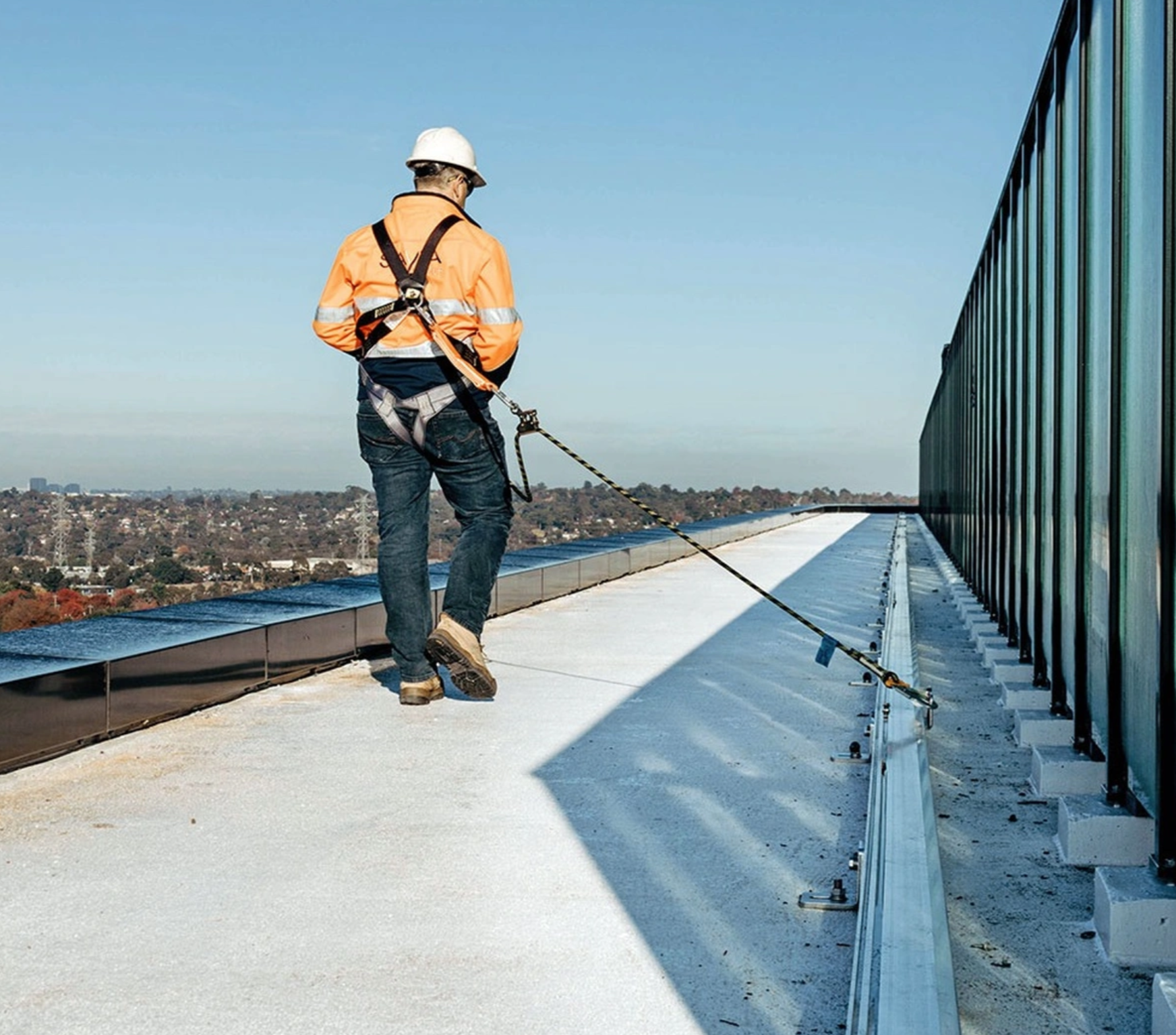 Worker in a harness and hi-vis connected by a lanyard to a rail system on a rooftop beside a glass balustrade